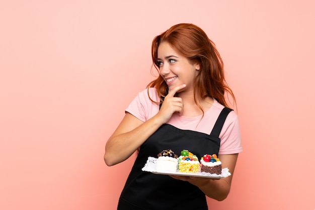 Ragazza della testarossa dell'adolescente che tiene i lotti di mini torte differenti sopra isolato pensando un'idea e guardando il lato