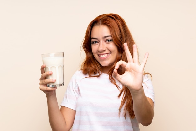 Ragazza della testarossa dell'adolescente che tiene un bicchiere di latte sopra isolato mostrando il segno giusto con le dita