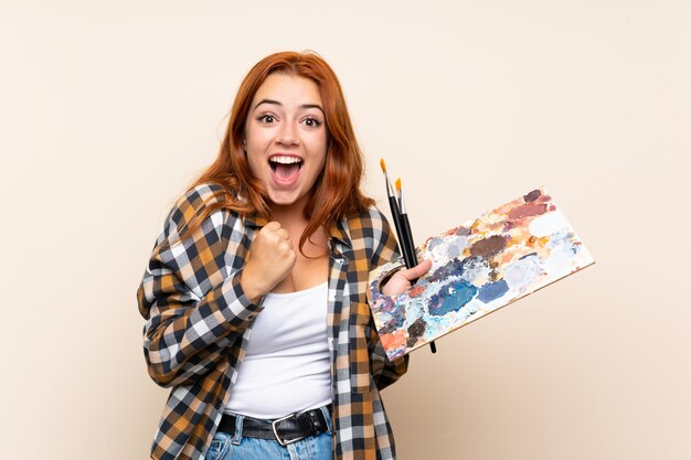 Ragazza della testarossa dell'adolescente che tiene una tavolozza sopra la parete isolata che celebra una vittoria