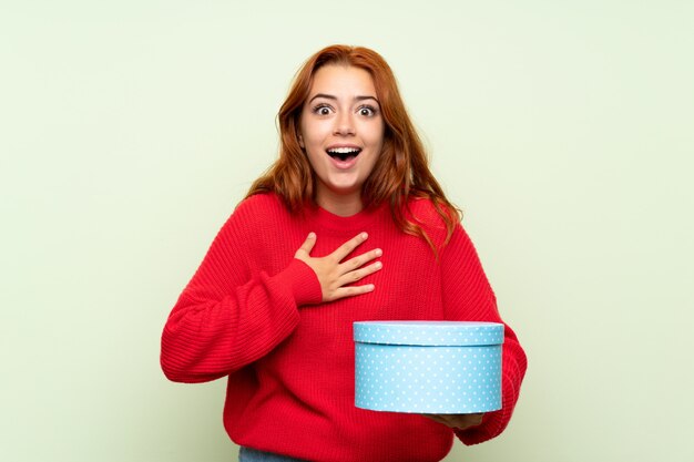 Ragazza della testarossa dell'adolescente con il maglione sopra il contenitore di regalo verde isolato della tenuta del fondo