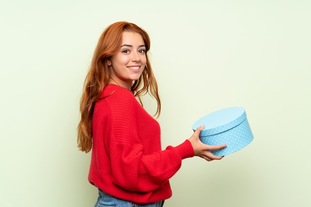 Ragazza della testarossa dell'adolescente con il maglione sopra il contenitore di regalo verde isolato della tenuta