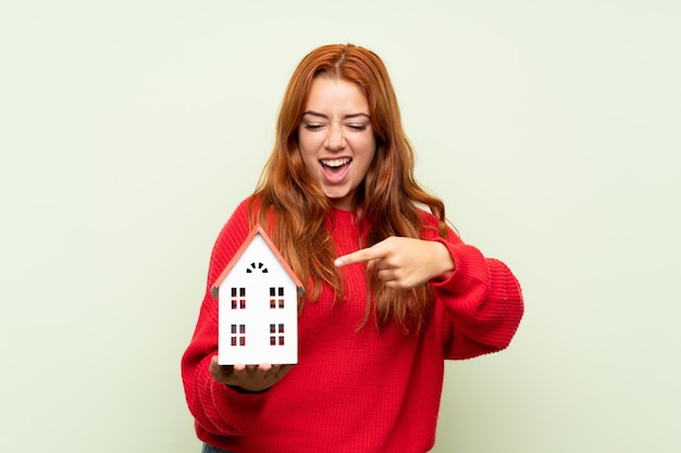 Ragazza della testarossa dell'adolescente con il maglione sopra la parete verde isolata che tiene una piccola casa