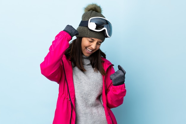 Ragazza dello sciatore della corsa mista con i vetri di snowboard sopra la parete blu isolata che celebra una vittoria