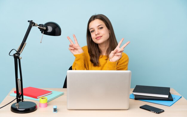 Ragazza dello studente che studia nella sua casa isolata sulla parete blu che mostra il segno di vittoria con entrambe le mani