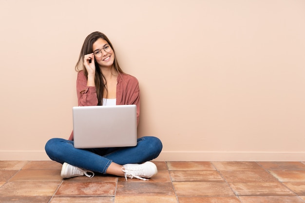 Ragazza dello studente dell'adolescente che si siede sul pavimento con un computer portatile con i vetri e felice