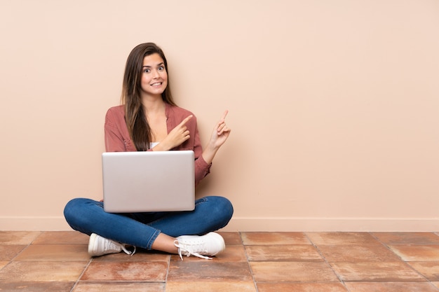 Ragazza dello studente dell'adolescente che si siede sul pavimento con un computer portatile spaventato e che indica il lato