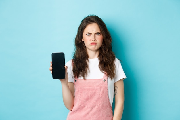 Ragazza delusa che si acciglia mentre mostra lo schermo vuoto dello smartphone, sembra dispiaciuta o arrabbiata, in piedi su sfondo blu.