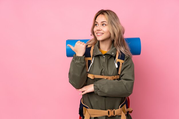 Ragazza di alpinista adolescente con un grande zaino isolato sul muro rosa che punta verso il lato per presentare un prodotto