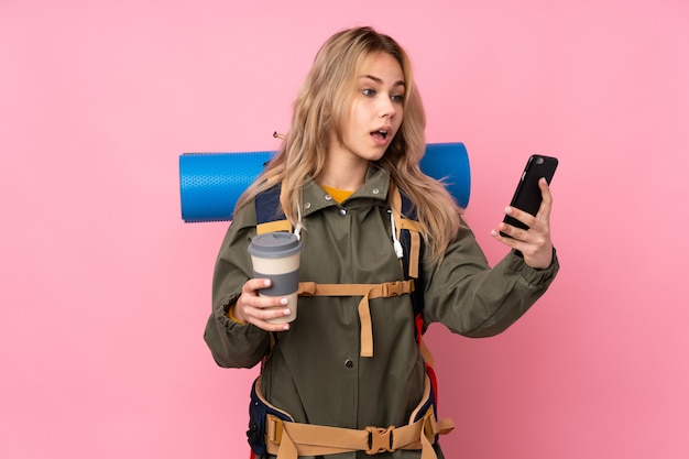 Ragazza di alpinista adolescente con un grande zaino sul muro rosa tenendo il caffè da portare via e un cellulare