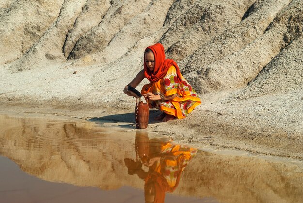 Ragazza di aspetto orientale in sari e hijab riempie la brocca d'acqua nella zona arida