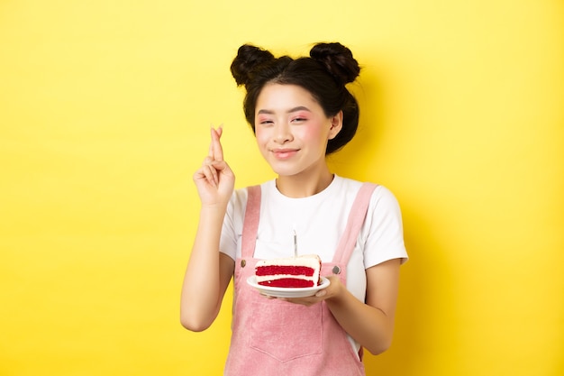 Ragazza di buon compleanno con trucco glamour, esprimere desideri e festeggiare, incrociare il dito per il sogno che si avvera, in piedi con la torta sul giallo.