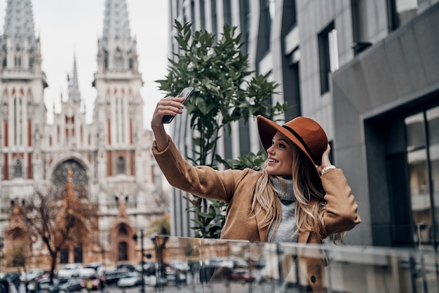 Ragazza di città con un grande stile. Attraente giovane donna sorridente con cappello e cappotto che si fa selfie mentre trascorre del tempo in città