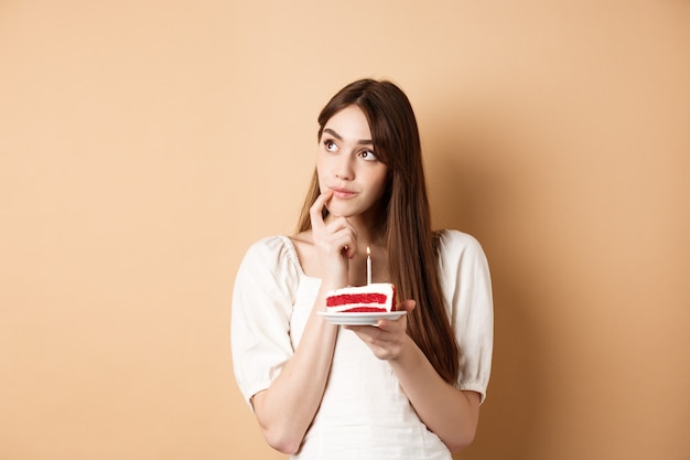 Ragazza di compleanno sognante guarda da parte e pensa al desiderio, soffiando la candela sulla torta, in piedi su sfondo beige.