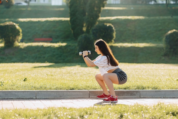 Ragazza di forma fisica che fa le esercitazioni con i dumbbells sul tramonto all&#39;erba fuori