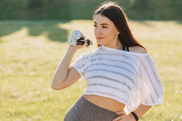 Ragazza di forma fisica che fa le esercitazioni con i dumbbells sul tramonto all&#39;erba fuori