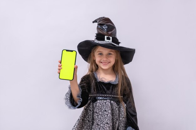 Ragazza di Halloween su sfondo grigio Bambino in costume da strega Banner di Halloween felice con spazio per la copia La ragazza tiene in mano lo smartphone con schermo verde Mock up Negozio Internet Acquisti online con le vendite