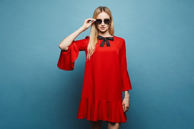 Ragazza di modello bionda bella, alla moda e sexy in occhiali da sole alla moda e in un breve abito rosso alla moda con fiocco nero in posa, isolato