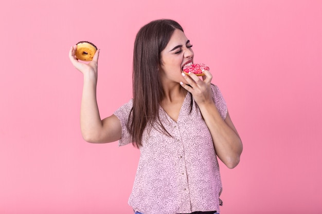 Ragazza di modello di bellezza che mangia ciambelle colorate. Donna in stile gioiosa divertente scegliendo dolci su sfondo rosa.