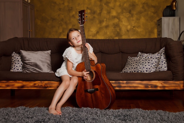 Ragazza di quattro anni abbastanza giovane con la chitarra che si siede sul divano all'interno della stanza. Bambino felice a casa