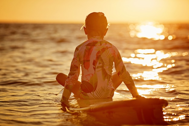 Ragazza di retrovisione che si siede su una tavola da surf durante il tramonto, mare calmo senza onde di luce arancione