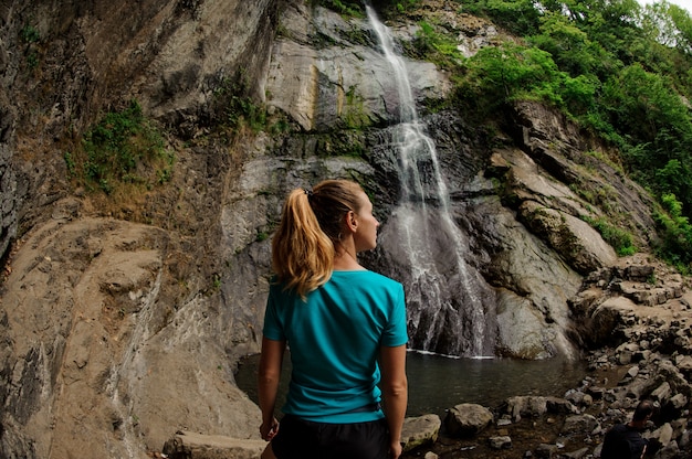 Ragazza di retrovisione della foto dell'occhio di pesce che sta e che esamina la bella cascata nella foresta