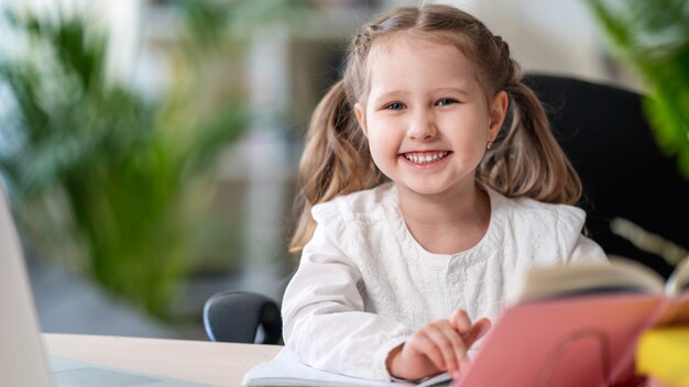 Ragazza di risata sveglia che si siede ad una tabella con un libro di testo. concetti di e-learning