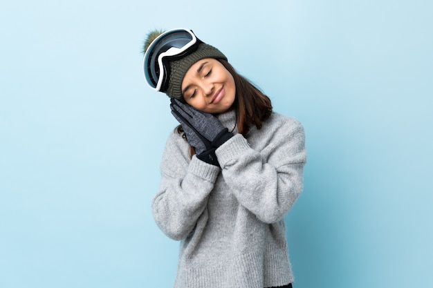 Ragazza di sciatore di razza mista con occhiali da snowboard su blu isolato che fa gesto di sonno nell'espressione dorable