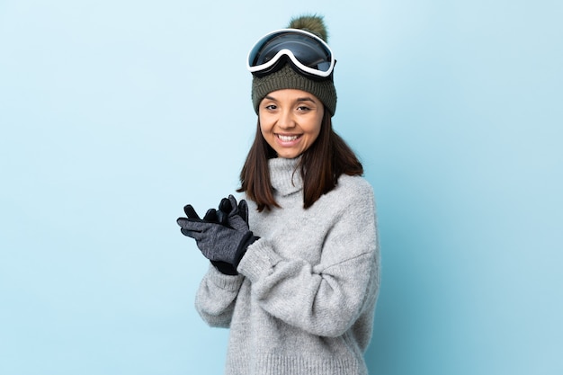 Ragazza di sciatore di razza mista con occhiali da snowboard su sfondo blu isolato che applaude.