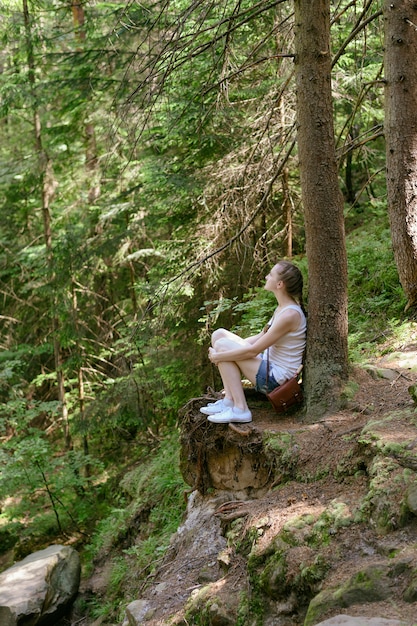 Ragazza di sogno che si siede su una pietra circondata dalla foresta di conifere un giorno soleggiato