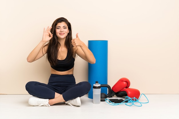 Ragazza di sport dell'adolescente che si siede sul pavimento che mostra segno e pollice giusti sul gesto