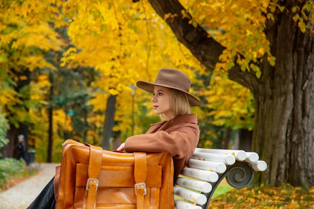 Ragazza di stile in cappotto marrone con la valigia che si siede su una panchina nel parco d'autunno