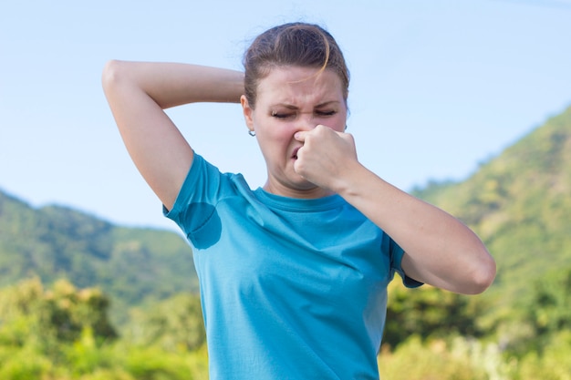 Ragazza disgustata, donna con maglietta sudata, ascelle bagnate e sporche, odore di sudore. Usando il deodorante, la signora dispiaciuta aggrotta le sopracciglia, si tappa il naso con le dita a causa dell'odore cattivo. Iperidrosi.