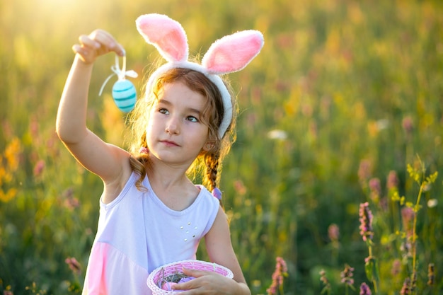 Ragazza divertente carina con uova di Pasqua dipinte in primavera in natura in un campo con luce solare dorata e fiori Vacanze di Pasqua Coniglietto pasquale con orecchie uova colorate in un cestino