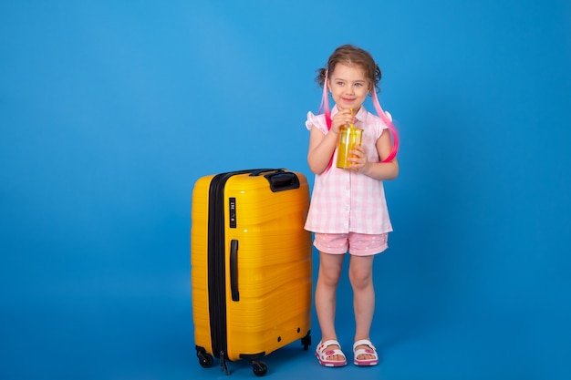 Ragazza divertente del bambino in vestiti rosa che tengono un bicchiere di succo d'arancia con la valigia gialla sulla superficie blu.
