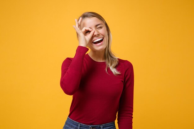 Ragazza divertente della giovane donna bionda in abbigliamento casual che posa isolata sul fondo giallo arancione della parete. Concetto di stile di vita delle persone. Simula lo spazio della copia. Tenendo la mano vicino all'occhio, imitando il binocolo o gli occhiali.