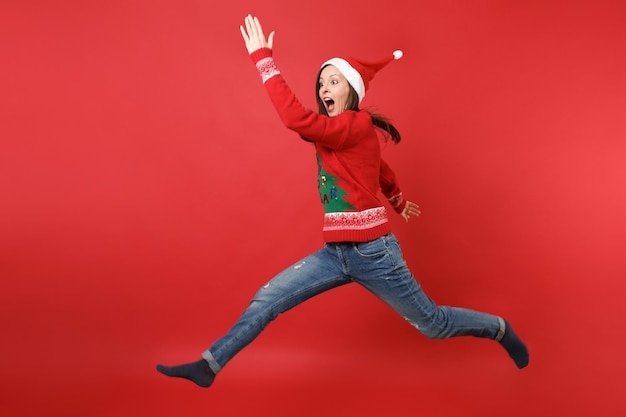 Ragazza divertente di Santa in maglione lavorato a maglia Cappello di Natale che salta, mani in aumento allargando le gambe, urlando isolato su sfondo rosso. Felice Anno Nuovo 2019 celebrazione festa concetto. Mock up copia spazio.