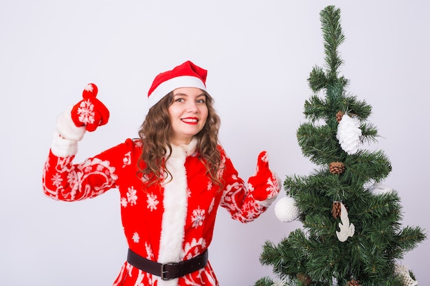 Ragazza divertente in costume da portare della santa di natale vicino all'albero di Natale che mostra i pollici aumenta il gesto.