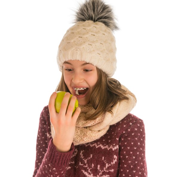 Ragazza divertente in maglione caldo, cappello, sciarpa e guanti che mangia una mela isolata su bianco