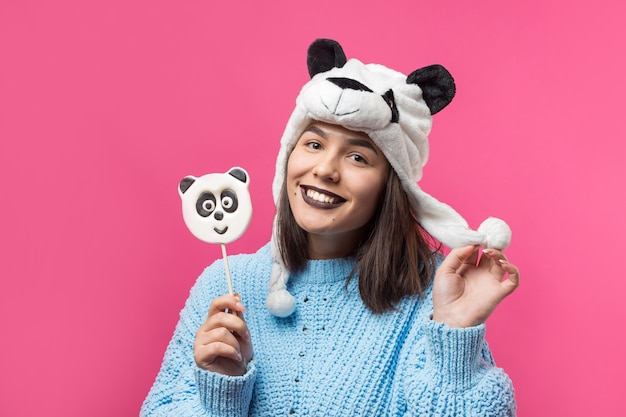 Ragazza divertente in piedi con un delizioso panda-lecca-lecca in mano e un cappello in testa su uno sfondo rosa.