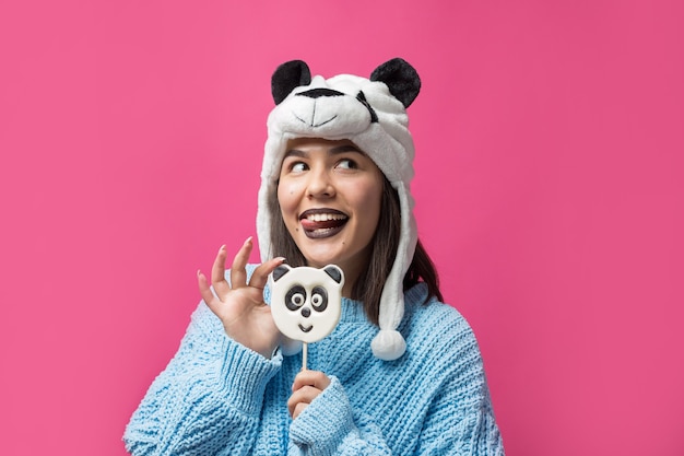 Ragazza divertente in piedi con un delizioso panda-lecca-lecca in mano e un cappello in testa su uno sfondo rosa.