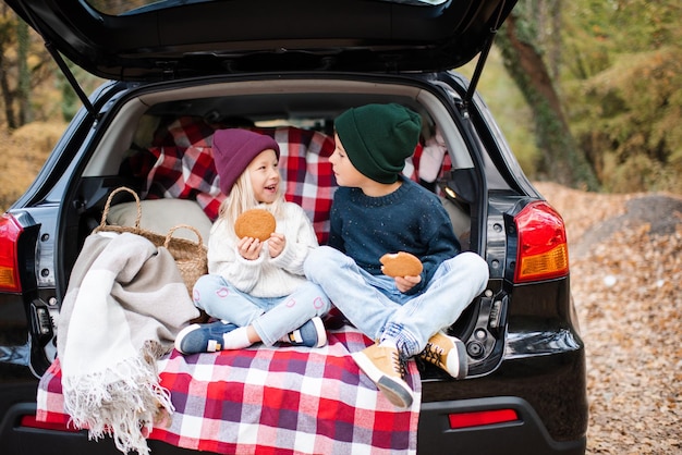 Ragazza e ragazzo sorridenti felici che mangiano biscotti con ragazza seduta in carrozzeria con coperta e decorazione all'aperto Infanzia