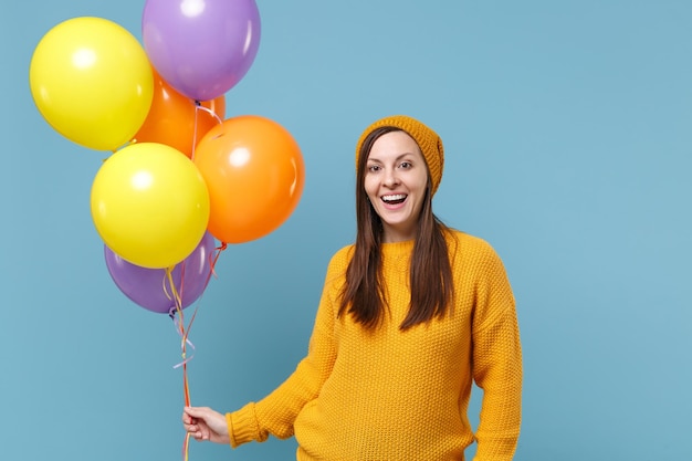 Ragazza eccitata della giovane donna in cappello del maglione che posa isolata sul ritratto dello studio del fondo blu. Festa di compleanno, concetto di emozioni della gente. Mock up spazio di copia. Festeggiando tenere palloncini colorati.