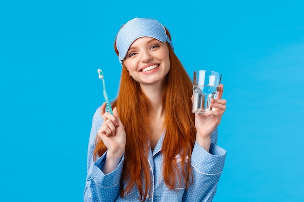 Ragazza entusiasta rossa sorridente e spensierata felice nella maschera di sonno, pigiama, tenendo acqua di vetro e spazzolino da denti, prendendosi cura dell'igiene della bocca, routine quotidiana dentale, parete blu