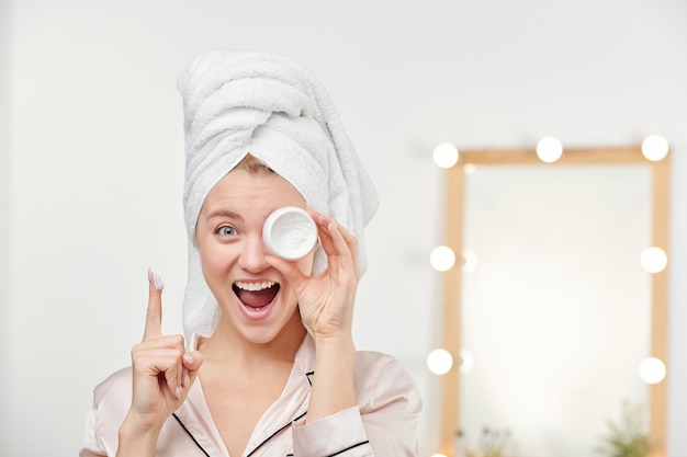 Ragazza estatica con un asciugamano bianco sulla testa che tiene il barattolo di crema idratante per il viso dall'occhio sinistro mantenendo l'indice sollevato