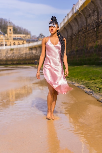 Ragazza etnica dominicana con trecce con un bel vestito rosa. La moda si gode l'estate passeggiando sulla spiaggia