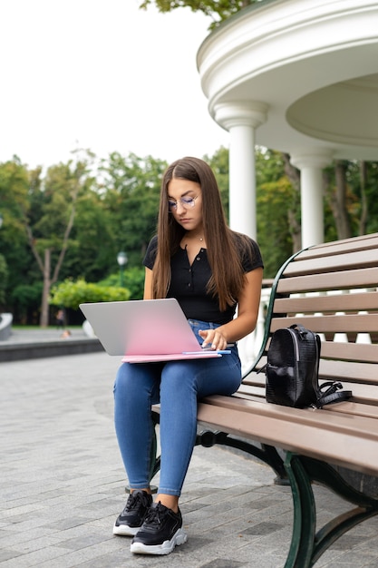 Ragazza europea dai capelli scuri seduta su una panchina del parco guardando un computer portatile con un notebook