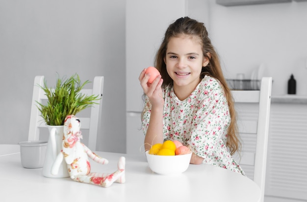 Ragazza europea di 10 anni in cucina al tavolo con uova di Pasqua rosa e gialle e un coniglio giocattolo