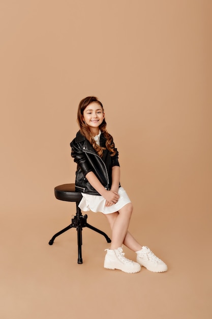 Ragazza fantastica. Integrale di una bambina divertente, a gambe incrociate e guardando con un sorriso, seduta su una sedia su un muro beige.