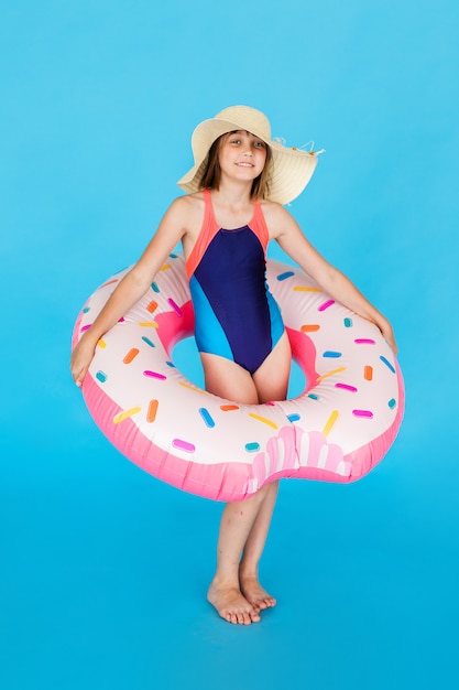Ragazza felice 10 anni con cappello di paglia in costume da bagno con anello di nuoto ciambella su una parete blu colorata