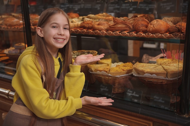 Ragazza felice adorabile che sorride, indicante all'esposizione di vendita al dettaglio del negozio di panetteria